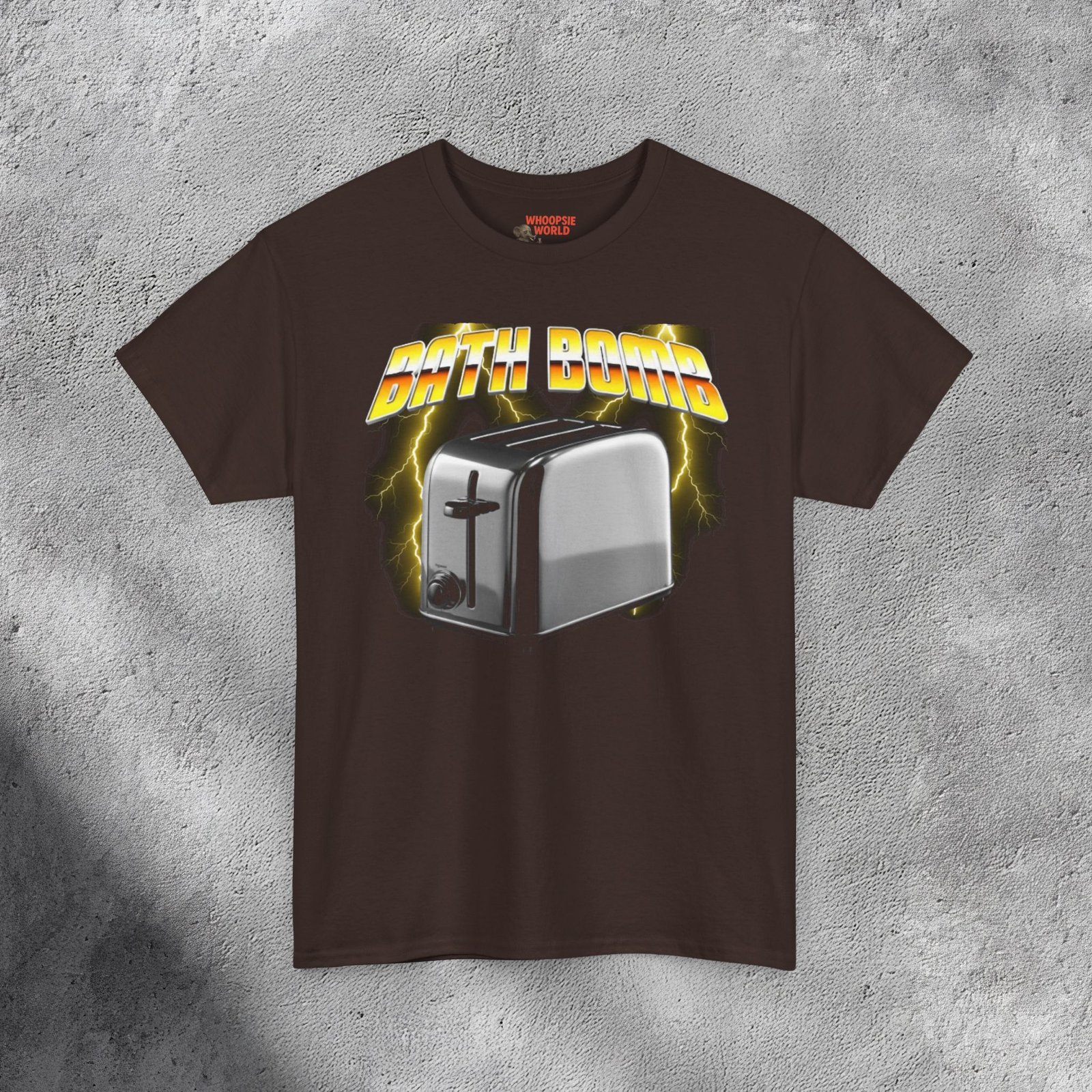 The T-shirt features a bold and humorous design showing a shiny silver toaster placed in the center, rendered in a photorealistic metallic style. Behind it, vivid yellow lightning bolts illuminate a smoky dark background, giving the image an electrifying energy. Above the toaster, the words “BATH BOMB” appear in large 3D yellow-and-orange gradient letters with a chrome outline, styled like a retro 1980s power-metal logo. The mix of humor, danger, and vintage aesthetic gives the shirt a striking and edgy appearance.