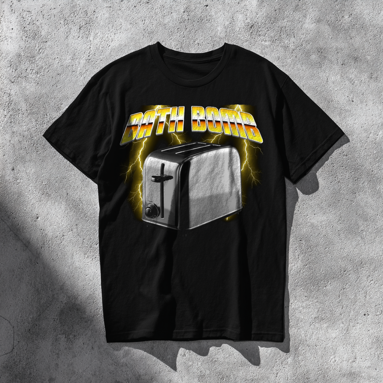 The T-shirt features a bold and humorous design showing a shiny silver toaster placed in the center, rendered in a photorealistic metallic style. Behind it, vivid yellow lightning bolts illuminate a smoky dark background, giving the image an electrifying energy. Above the toaster, the words “BATH BOMB” appear in large 3D yellow-and-orange gradient letters with a chrome outline, styled like a retro 1980s power-metal logo. The mix of humor, danger, and vintage aesthetic gives the shirt a striking and edgy appearance.